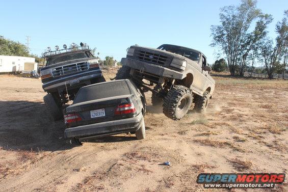 Two lifted trucks are crushing a black sedan in a dirt lot. One truck is on the right, the other on the left