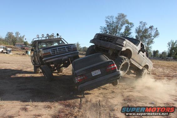 Two lifted Ford trucks on a dirt track, one crushing a black sedan