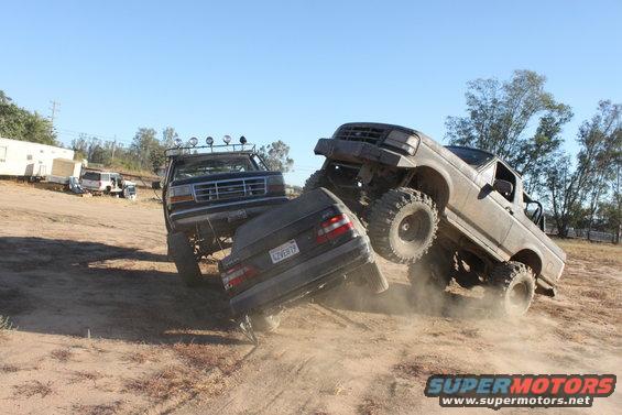 1992 Ford Bronco and another lifted truck crushing a sedan on a dirt lot
