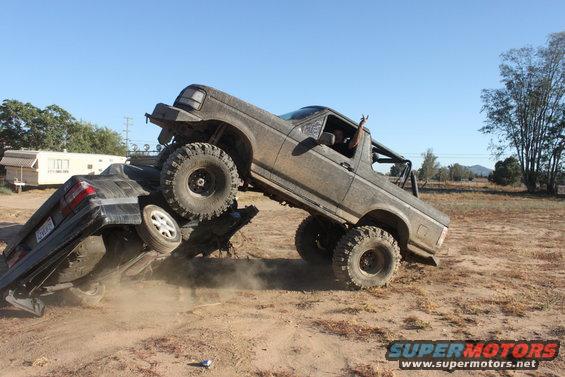 Mud-covered 1992 Ford Bronco on top of a crushed black sedan, with a person giving a peace sign from the driver's window