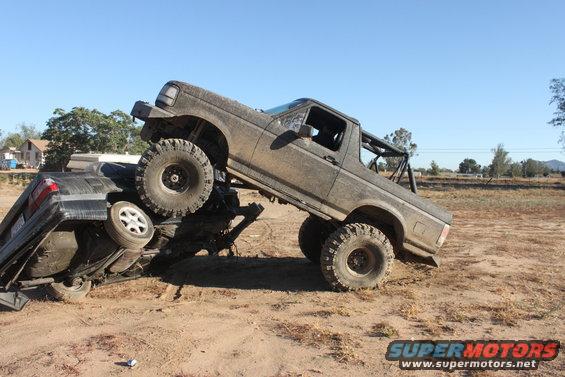 Muddy 1992 Ford Bronco on top of a crushed car, off-road scene