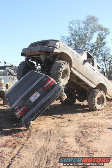 1992 Ford Bronco, lifted and modified, crushing a dark Volvo sedan on a dirt track