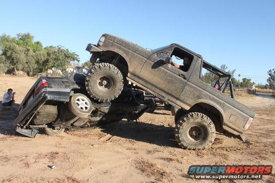 Mud-covered 1992 Ford Bronco on top of an overturned black car, driver visible inside the Bronco