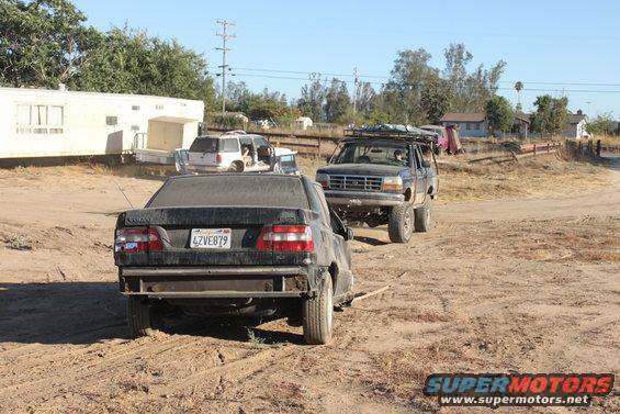 1992 Ford Bronco with roof rack and lifted suspension, followed by a damaged Volvo sedan on a dirt road