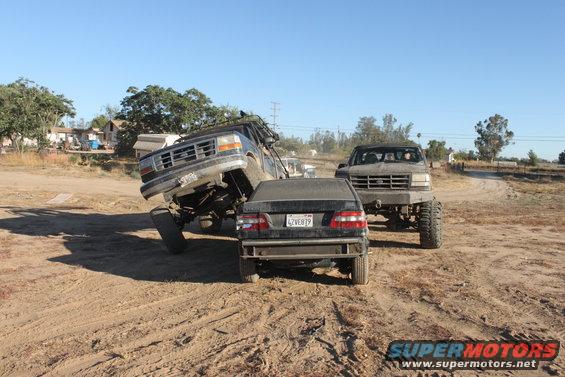 Two lifted trucks are in a dirt lot, one is on top of a sedan. Another truck is behind them