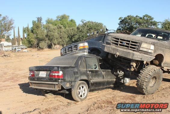 Lifted Ford truck on top of a wrecked sedan, both covered in dust. Truck grille visible