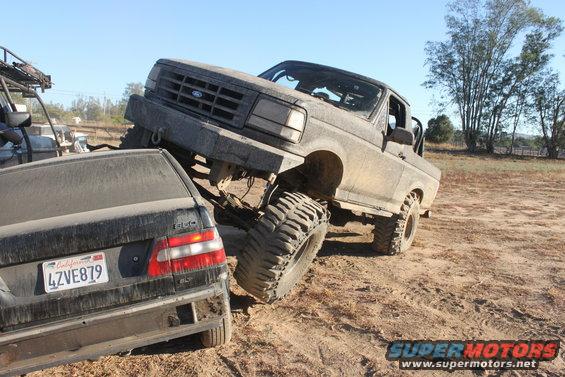 Mud-covered 1992 Ford Bronco on a dirt trail, front grille visible, with a black sedan underneath