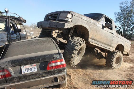 Lifted Ford truck driving over a black Volvo sedan on a dirt trail. Another vehicle is visible to the left