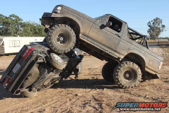Mud-covered 1992 Ford Bronco on top of an overturned black car, off-road tires, roll cage