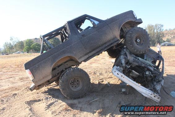 Black off-road truck on top of a crushed car, on a dirt hill