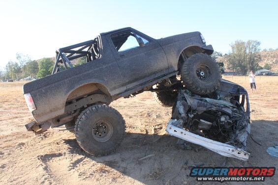 Black off-road Bronco on top of a crushed car, with a roll cage in the back. Person standing in background