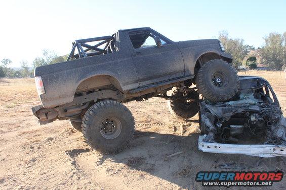 Black off-road truck with large tires on top of a crushed car. Roll cage visible in truck bed