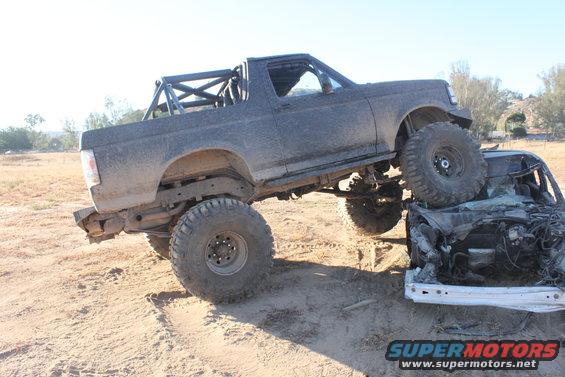 Dark gray 1992 Ford Bronco with a roll cage, lifted suspension, and large tires, crushing a wrecked white car