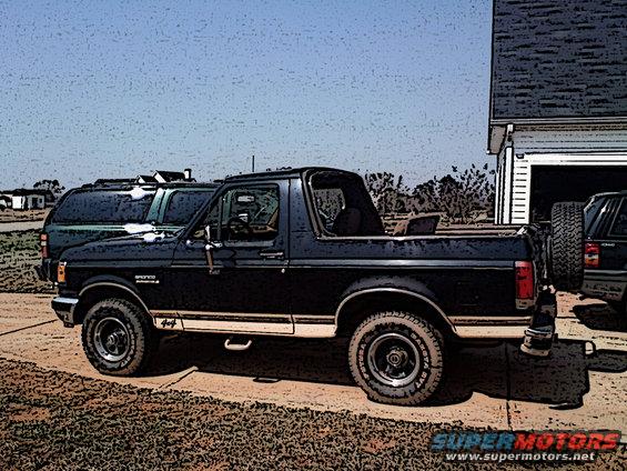 Dark blue 1990 Ford Bronco with tan stripe, parked next to a garage with a spare tire mounted on the rear of another vehicle
