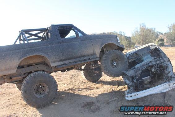 Dark gray 1992 Ford Bronco with large off-road tires crushing a white car