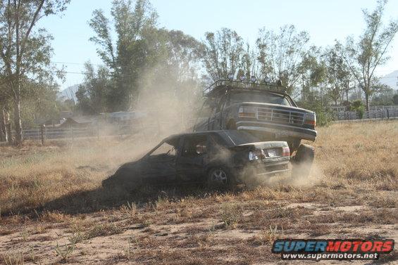 Large truck on top of a crushed sedan in a dusty field. The truck appears to be a 1992 Ford Bronco