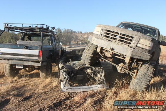 Two wrecked Ford trucks, one with a roof rack, are in a dry, grassy field under a clear blue sky