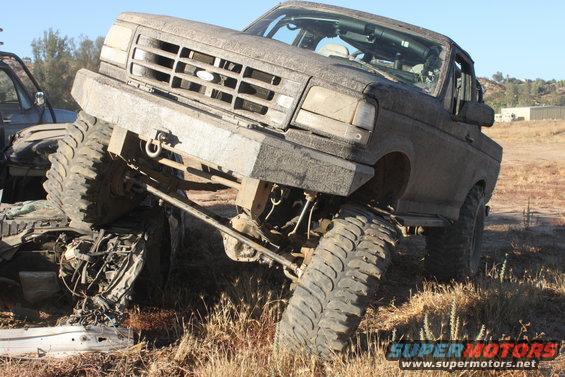 Muddy truck with large tires tilted on uneven ground. Another vehicle is partially visible to the left
