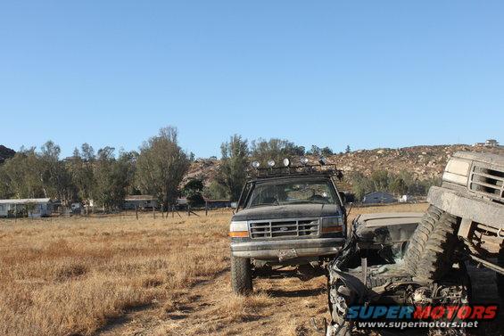 Black 1992 Ford Bronco with roof rack and lights parked next to a crushed vehicle