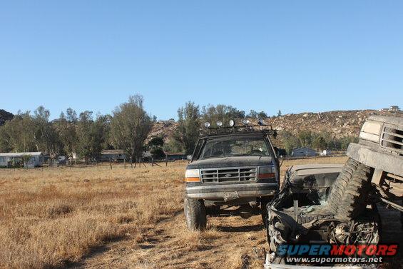 Black 1992 Ford Bronco with roof rack and lights, front view, next to a crushed vehicle