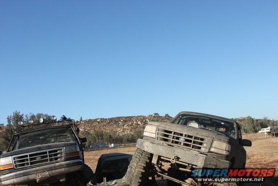 Two muddy trucks are parked on a dirt hill under a clear blue sky. One truck has extra lights on its roof