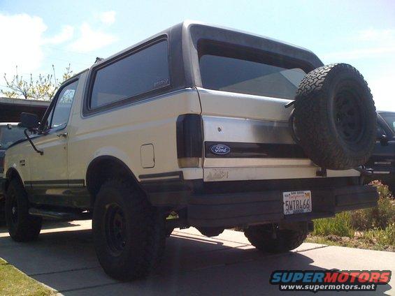 Cream-colored Ford Bronco with a black hardtop and spare tire mounted on the rear, viewed from the back-left