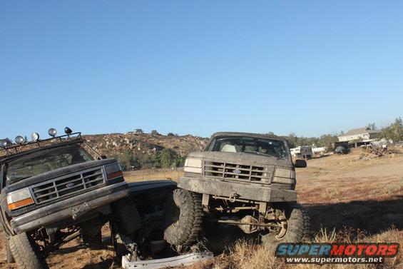 Two muddy Ford trucks, one with a roof rack and lights, are positioned at an angle on uneven terrain