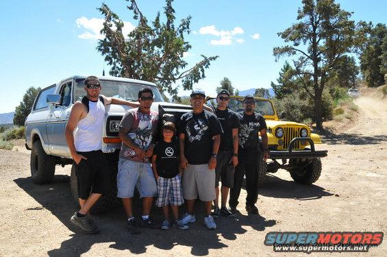 Group of guys and a kid posing in front of a light blue SUV and a yellow Jeep on a dirt road