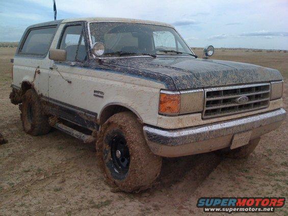 Mud-covered Ford Bronco parked in a dry, muddy field. The front grille and headlights are visible