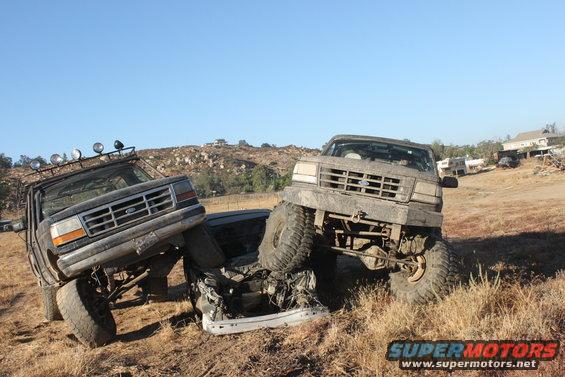 Two lifted Ford trucks, one on top of the other, stuck in dirt. The front truck is a 1992 Ford Bronco