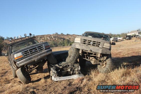 Two lifted Ford trucks are stuck in a field, one on top of a crushed car