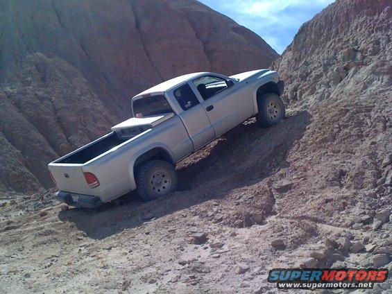 Silver pickup truck driving up a steep, rocky incline in a desert landscape. The truck has a bed cover