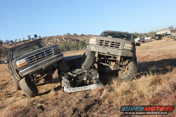 Two mud-covered 1992 Ford Broncos are positioned to crush a white car on a dry, grassy hillside