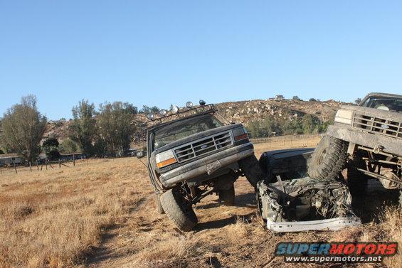 1992 Ford Bronco on its side, crushing a sedan, with another truck behind