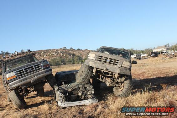 Two lifted trucks are on top of a crushed car in a dry, grassy field. One truck is dark, the other is lighter colored