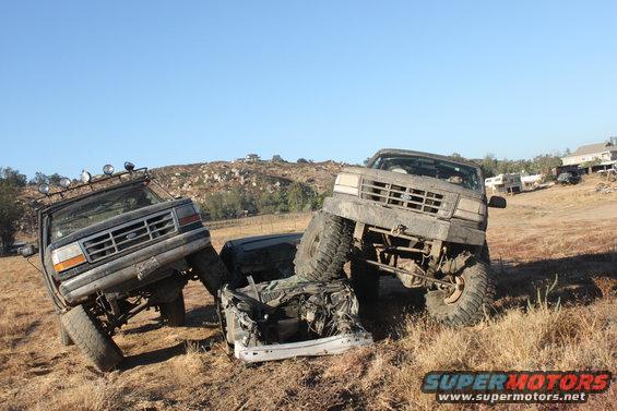 1992 Ford Bronco on the left, another Ford Bronco on the right, crushing a white car in a dry, grassy field