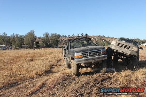 1992 Ford Bronco and another lifted truck on a dirt trail, with dry grass and hills in background