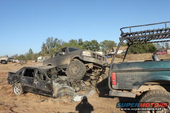Muddy trucks piled on top of a crushed car in a junkyard