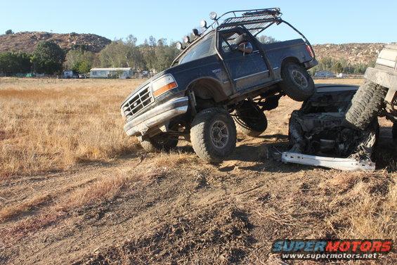 1992 Ford Bronco lifted high, front wheel on a crushed car. Another vehicle is partially visible on the right