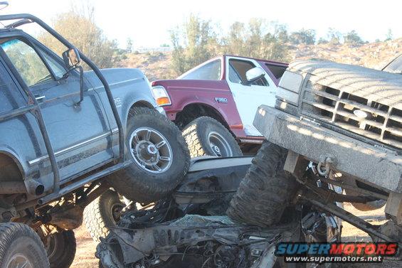 Several modified trucks are stacked on top of each other in a junkyard setting. One truck has a maroon cab and white bed