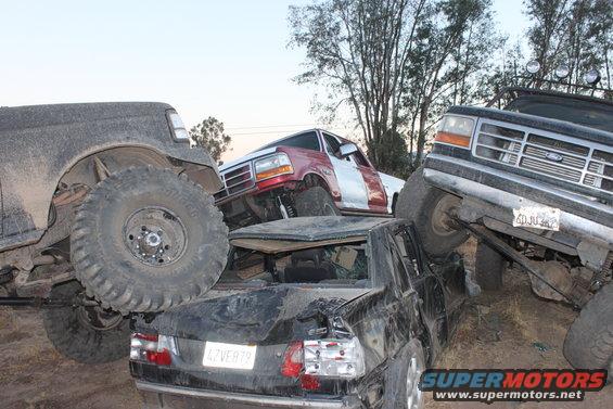 Three wrecked Ford trucks piled on top of each other, one with a red and silver two-tone paint job