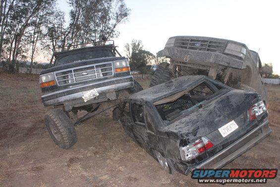 1992 Ford Bronco front end angled left, on top of a wrecked black sedan, with another vehicle behind