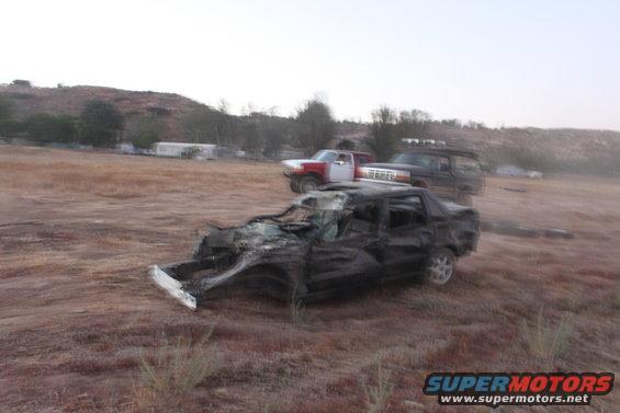 wrecked black car in foreground, red and white truck and dark SUV behind it in a dry field