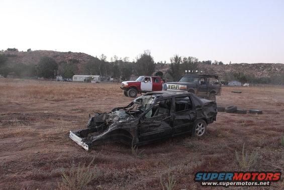 Wrecked black sedan in foreground, two trucks behind it on dry ground
