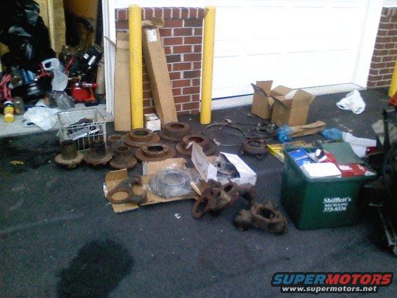 Pile of brake rotors, hubs, and other car parts on asphalt in front of a garage. Green recycling bin visible