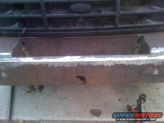 Custom steel bumper being fabricated in front of a vehicle grille. A jack is visible on the right