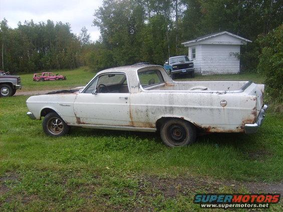 1966 Ford Ranchero pickup truck, white, rusty, parked in grassy area with other vehicles and a small building