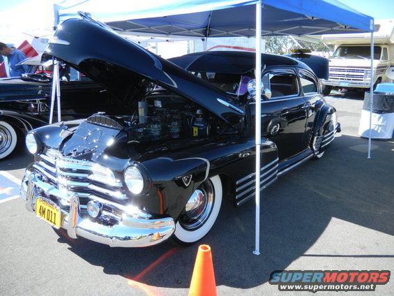 Black classic car with hood open, showing engine. Another vehicle and RV are visible in the background