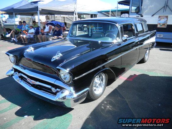 Black 1957 Chevrolet Nomad wagon parked outside with people sitting under tents in the background