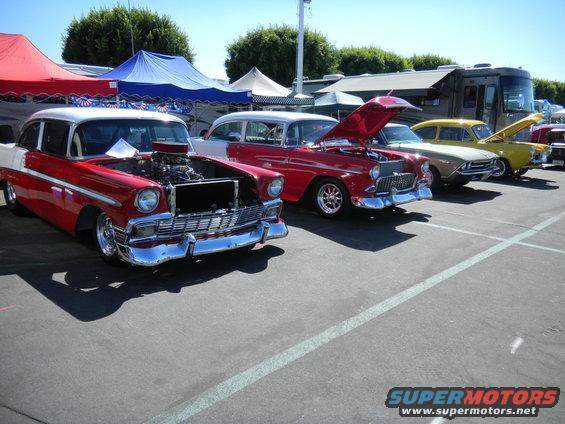 Several classic cars parked in a lot, some with hoods open. Tents and an RV are in the background